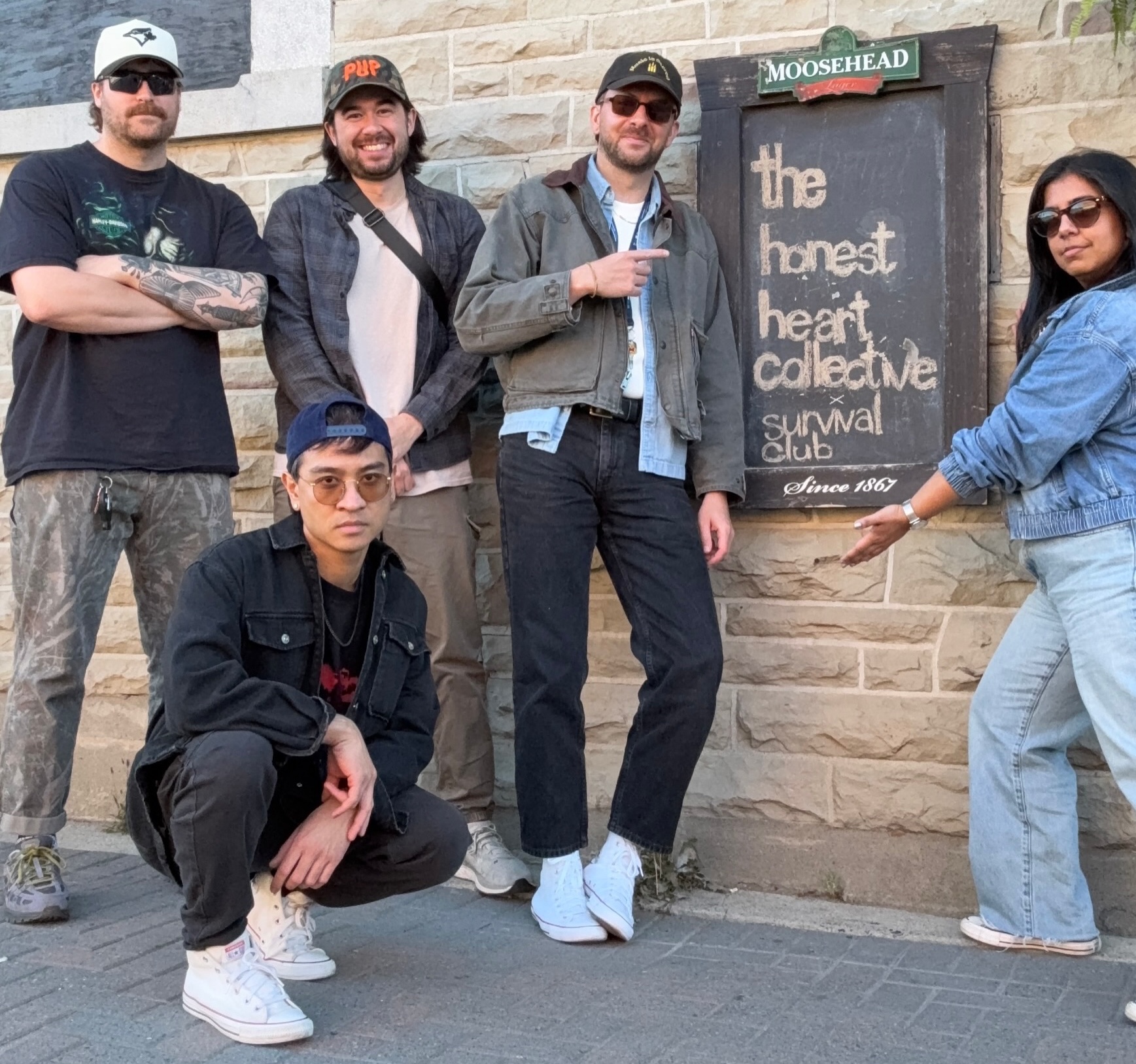 Survival Club posing in front of a bar sign that says The Honest Heart Collective x Survival Club